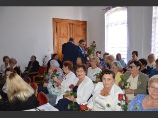 Inauguracja Roku Akademickego. Słuchacze Trzeciego Wieku siedzą na krzesłach, trzymają róże. Przy drzwiach Burmistrz Orzysza i Starosta Piski rozdają róże 