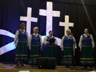 Na scenie kobiety z zespołu Orzyszanki. Przed nimi statywy mikrofonowe. W tle trzy krzyże.