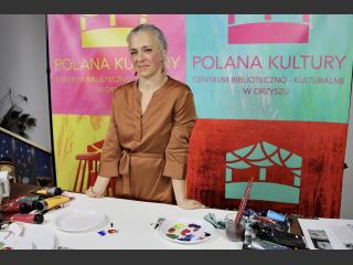 Monika Dąbkowska stoi przy stole. Na stole farby. Za nią logo Polanay Kultury w Orzyszu.