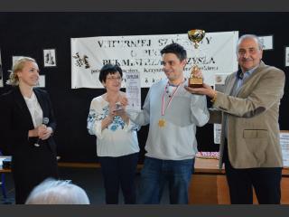 Mężczyzna, zwycięzca pierwszego miejsca w turnieju szachowym. W ręku trzyma puchar i dyplom, na szyji ma medal. Z prawej strony Wicestarosta Piski, z lewej strony Pani Dyrektor Polany Kultury.