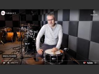 Zrzut ekranu z filmu na YouTube, na kanale Domu Kultury. Krzysztof Roszko instruktor gry na perkusji, siedzi, przed nim bęben, za nim perkusja.
