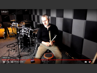 Zrzut ekranu z filmu na YouTube, na kanale Domu Kultury. Krzysztof Roszko instruktor gry na perkusji, siedzi, trzyma w ręku pałki do perkusji. Za nim perkusja. W studiu nagrań