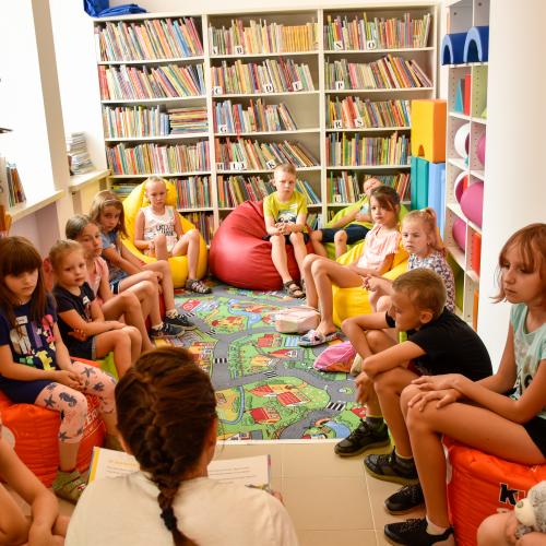 Dzieci w kąciuku dziecięcym w bibliotece. Przy ścianach regały z książkami.