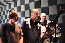 Trzech mężczyzn w studiu nagrań, przed nimi mikrofon.