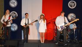 Zespół na scenie gra na gitarach i perkusji. Po środku dziewczyna trzyma mikrofon, śpiewa. W tle flaga Polski. Przegląd Piosenki Patriotycznej i Żołnierskiej.