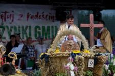 Msza odprawiana na scenie podczas dożynek gminnych w Rostkach Skomackich. Na scenie ksiądz, obok niego krzyż. Przed nim wieniec dożynkowy z chlebem.
