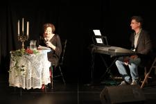Ewa Dałkowska siedzi przy stoliku z mikrofonem w ręku. Z prawej strony Rafał Gajewski przy instrumencie klawiszowym.
