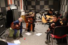 Grupka 4 gitarzystów siedzi na krzesłach w studio nagrań z gitarami w ręku.