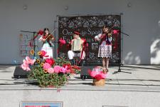Trójka dzieci w strojach ludowych na scenie podczas występu. Dwie dziewczynki z prawej i lewej grają na skrzypcach i jeden chlopiec po środku gra na akordeonie. Przed nimi statywy mikrofonowe ozdobione kwiatami i wstążkami.