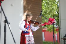 Na scenie dziewczynka w stroju ludowym gra na skrzypcach. Przed nią statyw mikrofonowy ozdobiony kwiatem. W tle drzewa.