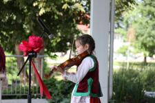 Dziewczynka w stroju ludowym, na scenie gra na skrzypcach. Przed nią statyw mikrofonowy. W tle drzewa.