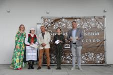 Na scenie Joanna Kamieniecka - Dyrektor Polany Kultury w Orzyszu, jury, oraz Pani Apolonia Nowak w stroju ludowym. Za nimi baner z wydarzenia.