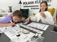 Dwie dziewczynki przy stole, na stole puzzle do kodowania. Za nimi na ścianie napis Polana Kultury.