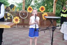 Dziewczynka w stroju ludowym, na scenie, gra na klarnecie. W tle dekoracyjne słoneczniki.