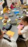 Dzieci przy stole podczas robienia laurek dla seniorów. Na stole kredki, liście, kolorowe kartki, wycinanki.
