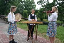 Trójka młodzierzy w strojach ludowych gra na instrumentach. Dziewczyna z lewej strony gra na tamburynie z bębenkiem, chłopak po środku gra na cymbałach, dziewczyna po prawej gra na skrzypcach.