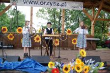 Trójka w strojach ludowcyh gra na instrumentach. Dziewczyna z lewej strony gra na tamburynie z bębenkiem, chłopak po środku gra na cymbałach, dziewczyna po prawej gra na skrzypcach. W tle dekoracje 