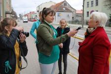 Młodzież przeprowdza wywiad z kobietą na ulicach Orzysza. Jedna z dziewczyn ma słuchwaki i trzyma mikrofon, dziewczyna z lewej robi zdjęcia aparatem, za nimi dziewczyna 