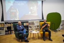 Dwóch mężczyzn siedzi na fotelach. W bibliotece Polany Kultury. Z prawej strony Artur Olchowy. Z tyłu wyświetlana prezentacja.