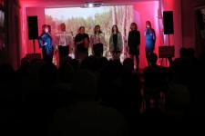 Na scenie część artystów tworzących płytę "Leć głosie po rosie". Z prawej strony pięć kobiet, z lewej strony dwóch mężczyzn i kobieta. W tle wyświetlana prezentacja multimedialna.