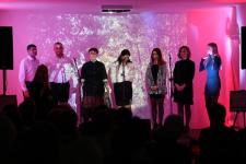 Na scenie część artystów tworzących płytę "Leć głosie po rosie". Z prawej strony pięć kobiet, z lewej strony dwóch mężczyzn. W tle wyświetlana prezentacja multimedialna. 