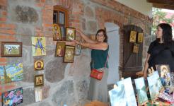 Budynek Muzeum Muzeum Michała Kajki w Ogródku. Na ścianach obrazy i ikony. Kobieta trzyma jedną z ikon i uśmiecha się.
